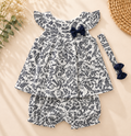 A baby girl's three-piece outfit set featuring a white and navy blue fern pattern, consisting of a ruffle-shoulder top with a navy bow, matching bloomers, and a long-sleeve top with a navy bow, laid flat on a beige background with decorative props.