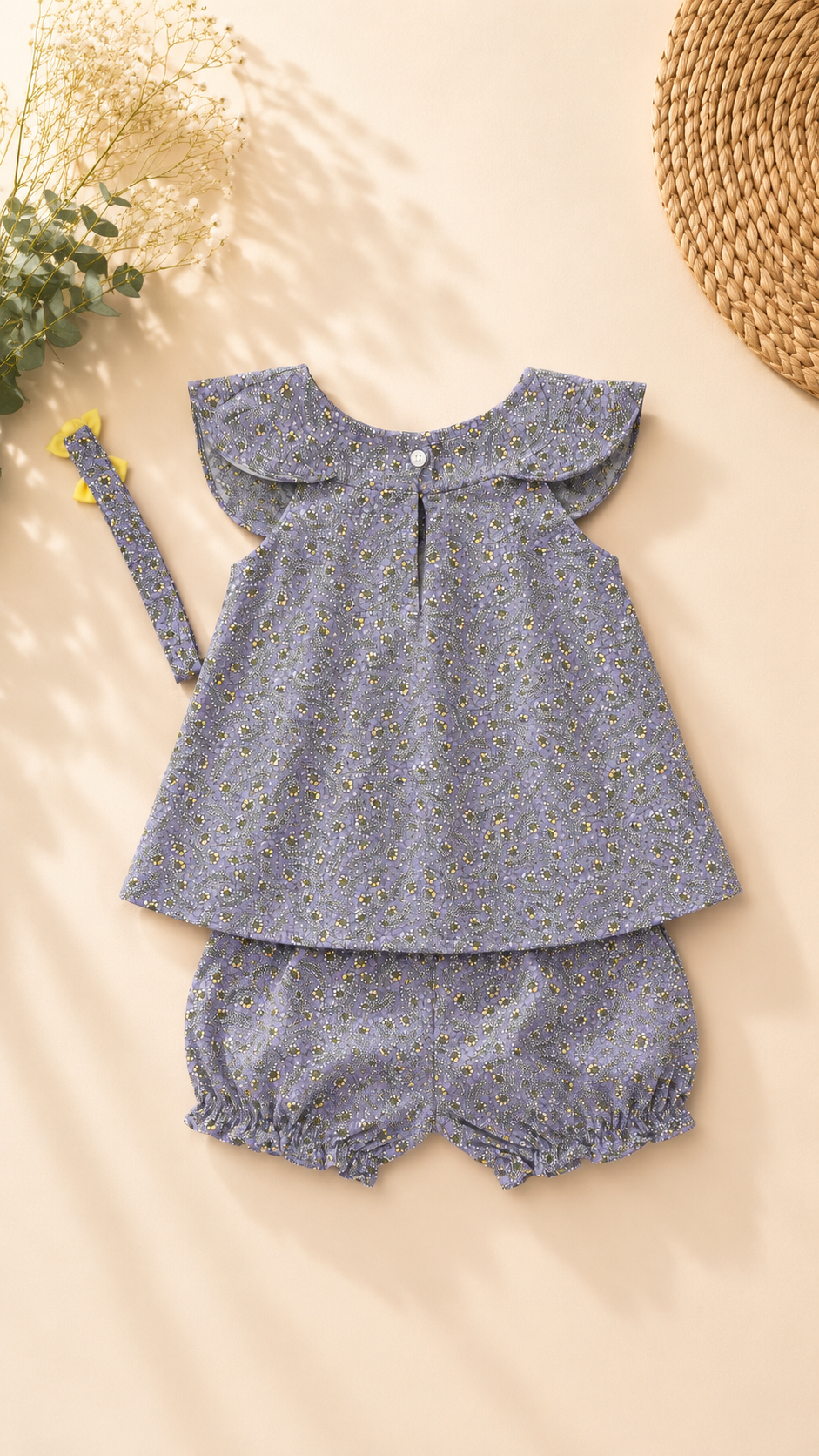 A baby girl's two-piece outfit set in a dusty blue-gray floral print, featuring a short-sleeve top with a ruffled collar and matching bloomers, laid flat on a beige background with a coordinating headband and decorative props.