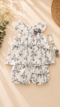 A baby girl's two-piece outfit set featuring a white short-sleeve romper and matching shorts with a delicate black floral print, styled with a gray bow on the bodice and a matching headband. The set is laid flat on a beige background with decorative dried flowers and a woven straw hat.