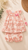 A baby girl's two-piece outfit set featuring a ruffled sleeveless top and matching bloomers in a pink and white toile pattern, styled with a pink bow headband on a beige background with decorative dried flowers and a woven straw hat.