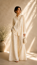 A woman stands in a sunlit studio wearing a matching cream-colored embroidered shirt and wide-leg pants set, holding a small woven handbag. The background features a textured beige wall with soft leaf shadows.
