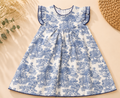 A sleeveless baby dress with a blue and white toile de Jouy pattern, featuring ruffled shoulder straps and a scalloped blue hem, laid flat on a beige background with decorative dried flowers and a woven mat.
