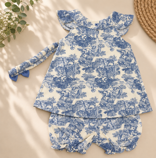 A baby girl's two-piece outfit set featuring a long-sleeve ruffle-sleeve top and matching bloomers, both in a blue and white toile de Jouy pattern, laid flat on a beige background with decorative props.