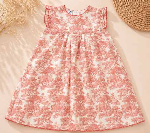 A sleeveless baby dress with a pink and white toile de Jouy pattern, laid flat on a beige background with dried flowers and a woven straw hat as props.
