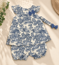 A baby girl's two-piece clothing set featuring a long-sleeve top and matching shorts in a blue and white toile de Jouy pattern, styled flat-lay on a beige background with decorative greenery.
