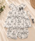 A baby girl's two-piece outfit set featuring a white ruffle-sleeve top and matching bubble shorts with a black floral print, styled with a gray bow and a matching headband on a beige fabric background.