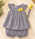 A baby girl's two-piece outfit set featuring a short-sleeve top and matching bloomers in a lavender-gray floral print, decorated with yellow bows, shown with a matching headband on a beige background with decorative dried flowers and a woven hat.