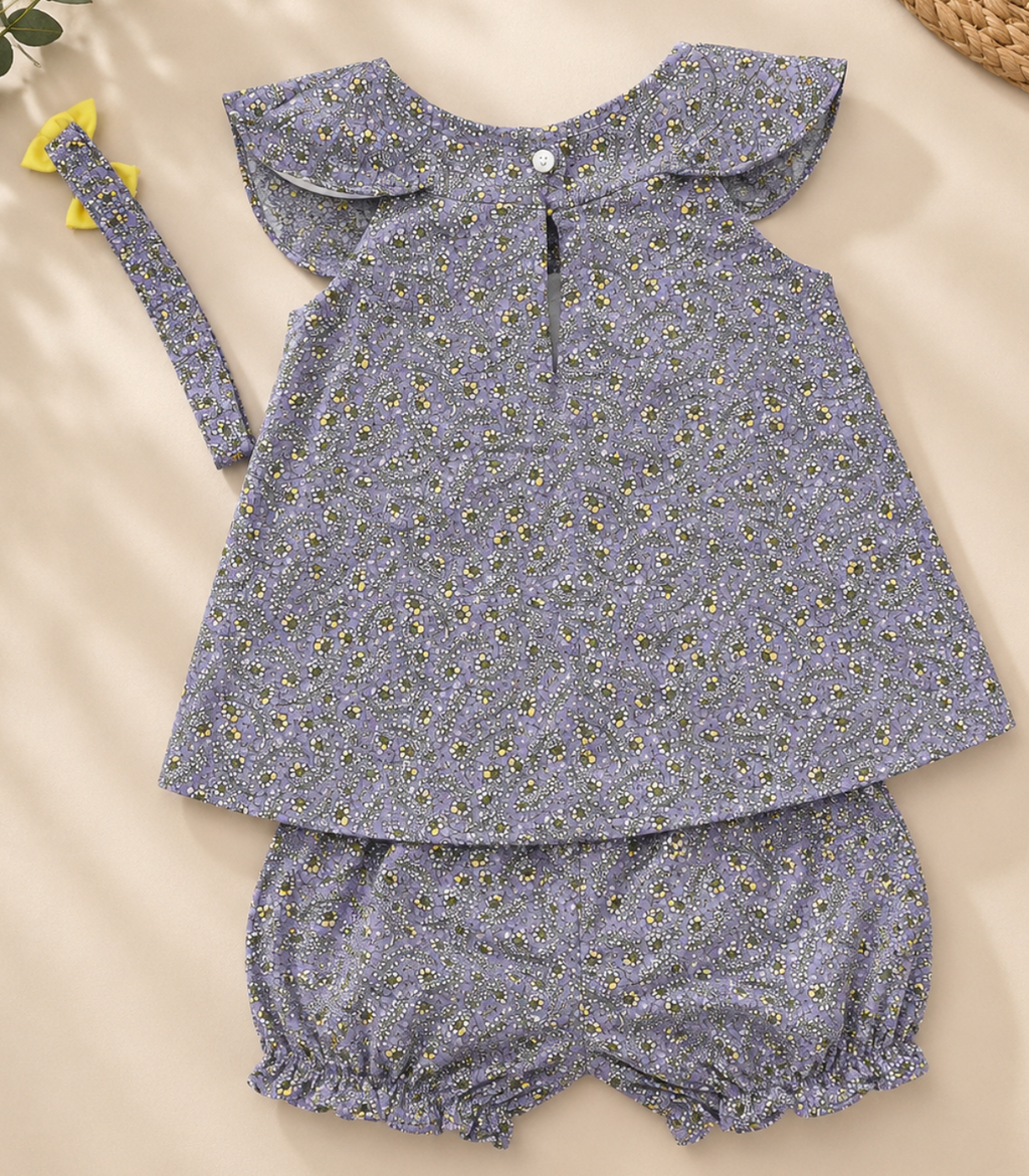 A baby girl's three-piece outfit set featuring a short-sleeve top with a ruffled Peter Pan collar, matching bloomers, and a headband, all in a dusty lavender-blue floral print with tiny yellow and white flowers. The set is laid flat on a beige background with decorative dried flowers and a woven basket corner.