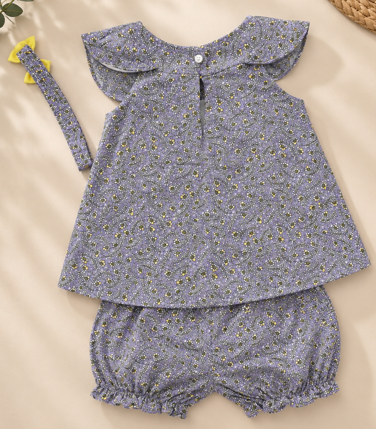 A baby girl's three-piece outfit set featuring a short-sleeve top with a ruffled Peter Pan collar, matching bloomers, and a headband, all in a dusty lavender-blue floral print with tiny yellow and white flowers. The set is laid flat on a beige background with decorative dried flowers and a woven basket corner.