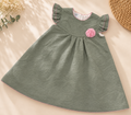 A toddler girl's sage green textured dress with ruffled cap sleeves, a gathered neckline, and a pink fabric rose applique on the chest. The dress is laid flat on a beige background decorated with dried flowers and a woven straw hat.
