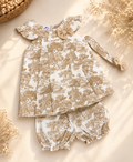 A baby girl's two-piece outfit set featuring a ruffled long-sleeve top and matching bloomers, both in a beige and white toile pattern. The set is styled in a flat lay with dried flowers and a woven basket on a warm beige background.