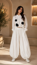 A woman modeling a white long-sleeve button-up shirt and matching wide-leg harem pants set, featuring black circular patch details on the chest, in a warm, sunlit indoor setting.