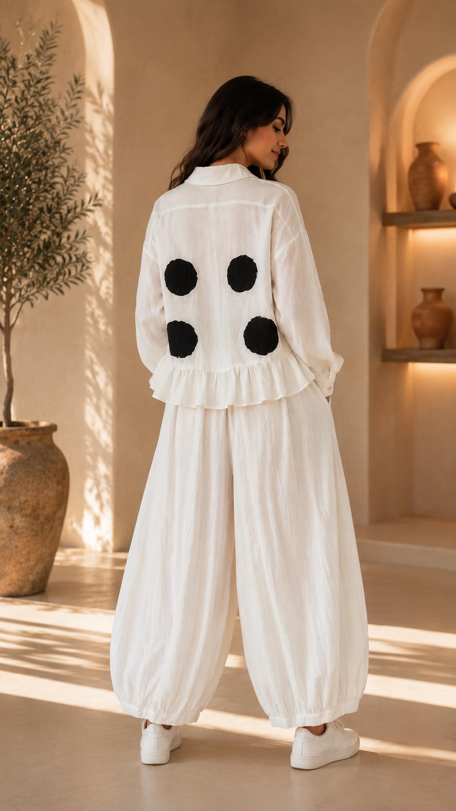 A woman stands with her back to the camera wearing a white long-sleeve blouse with four black circular patches on the back and matching white wide-leg pants, in a warm, sunlit interior with beige walls and arches.