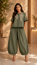 A woman modeling an olive green two-piece outfit consisting of a short-sleeve button-up top with floral embroidery on the chest and matching wide-leg harem pants, set against a warm, sunlit indoor background with potted plants.